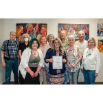 group of 10 people standing in two rows in an art gallery with colorful paintings behind them. Woman in center in front is holding a certificate.
